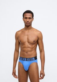 Μπλε σλιπ Calvin Klein με μαύρη ζώνη που διαθέτει λευκό κείμενο λογότυπου. Το ύφασμα φαίνεται λείο και εφαρμοστό, σχεδιασμένο για να έχει σφιχτό σχήμα.