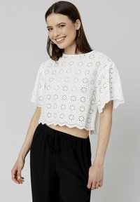 Top court blanc avec broderie ajourée à motifs floraux, bords festonnés et manches courtes, associé à un pantalon noir à cordon de serrage.