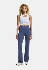 Biały krótki top na ramiączkach z wycięciem, połączony z spodniami w ciemnym niebieskim kolorze o wysokim stanie. Modelka stoi w białych tenisówkach.