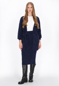 Navigatie gebreide cardigan met wijde mouwen en bijpassende rok, beide van zacht, textuurstof. Zwarte kniehoge laarzen met gespsdetails.