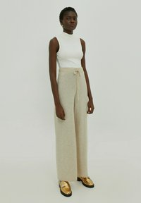 Beige weite Hosen mit einer weichen Stricktextur und elastischem Bund, kombiniert mit einem figurbetonten weißen ärmellosen Oberteil und glänzenden goldfarbenen Loafers.