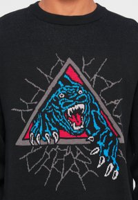 Zwarte trui met een blauwe tijger die uit een grijs en rood driehoekig kader springt, met uitgeslagen klauwen en open mond die scherpe tanden laat zien.