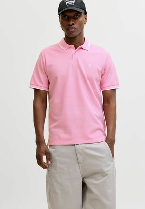 Vyras, dėvintis rožinę trumpomis rankovėmis polo marškinėlį, šviesiai smėlio spalvos kelnes ir juodą kepuraitę, stovintis priešais paprastą šviesią foną.