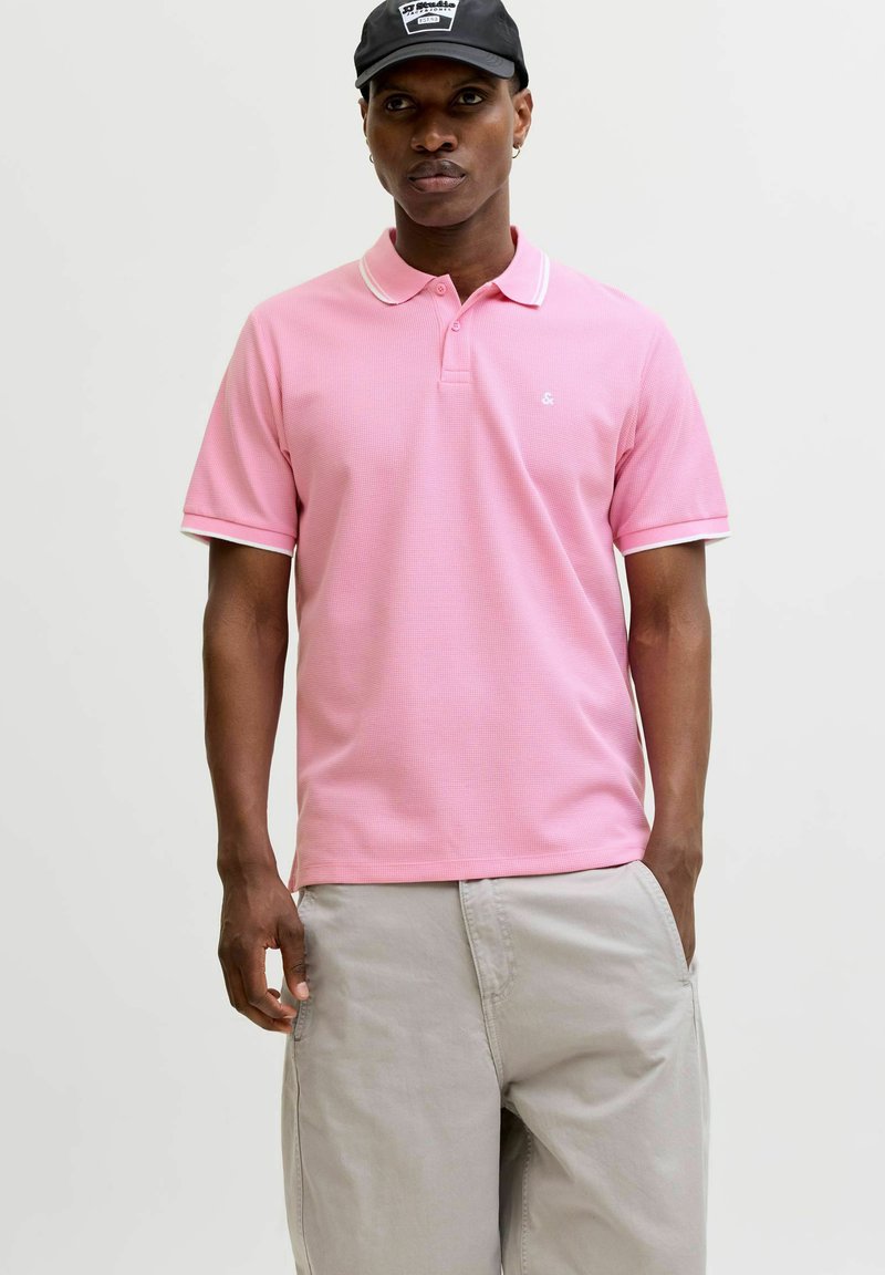 Mees, kes kannab roosat lühikeste varrukatega polo särki, heledaid beeži pükse ja musta nokamütsi, seisab ühtlase heleda tausta ees.