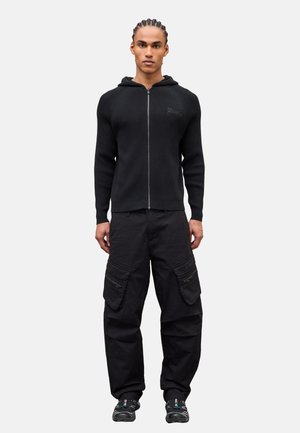 Mann mit geflochtenen Haaren steht mit dem Gesicht nach vorne und trägt einen schwarzen Hoodie mit Reißverschluss, schwarze Cargohosen mit Taschen und schwarze Sneaker.