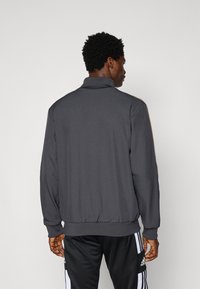 Σκούρο γκρι μπουφάν με φερμουάρ και ψηλό γιακά, ριμπ τελειώματα στα μανίκια και τη μέση, λεία υφή και ρίγες στο πλάι. Φορεμένο πάνω από μαύρο αθλητικό παντελόνι.