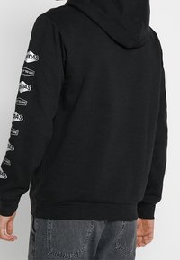 Osoba v černé mikině s kapucí a bílými textovými logy na levém rukávu, s vybledlými černými džíny, pohled zezadu.