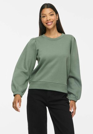 Kvinde iført en grøn langærmet puffet sweatshirt og sorte bukser, smilende med hænderne afslappet ved siderne, mod en ensfarvet baggrund.