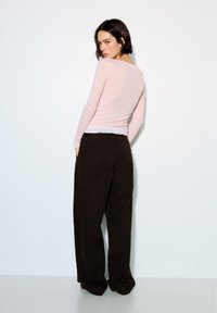 Frau mit kurzen dunklen Haaren, die ein hellrosa langärmeliges Oberteil und lockere schwarze Hosen trägt, steht vor einer weißen Wand und schaut zurück.