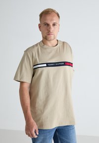 Beige puuvillane t-särk lühikeste varrukatega, millel on horisontaalne musta, valge ja punase triibuga logo riba rinnal.