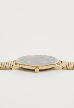 Montre dorée avec un cadran noir, bracelet en maille métallique, design minimaliste et boîtier rond. Présente des repères horaires et une couronne sur le côté.