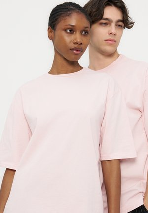 Γυναίκα και άνδρας στέκονται κοντά, φορούν και οι δύο απλά ροζ ανοιχτά φαρδιά μπλουζάκια μπροστά σε λευκό φόντο.