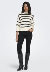 Cream and black striped sweater with a high collar, ribbed texture, wide sleeves, paired with black skinny jeans and knee-high black boots.