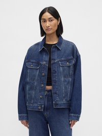 Denimjakke i mørk blå, med en avslappet passform, store lommer, knappelukking og subtil falming. Kombinert med en svart topp.