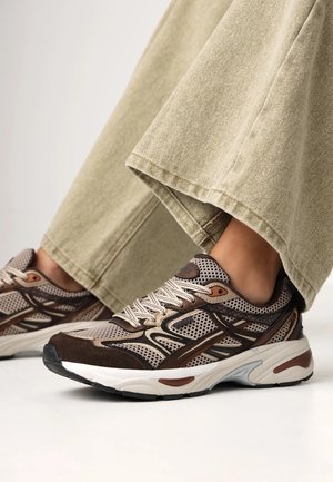 Person trägt beige Hosen mit weitem Bein und braun-beige Sportschuhe und steht auf einer weißen Oberfläche.