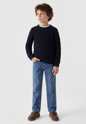 Dreng med krøllet hår står med den ene hånd i lommen iført mørk strikket sweater, blå jeans og brune sko mod en ensfarvet baggrund.