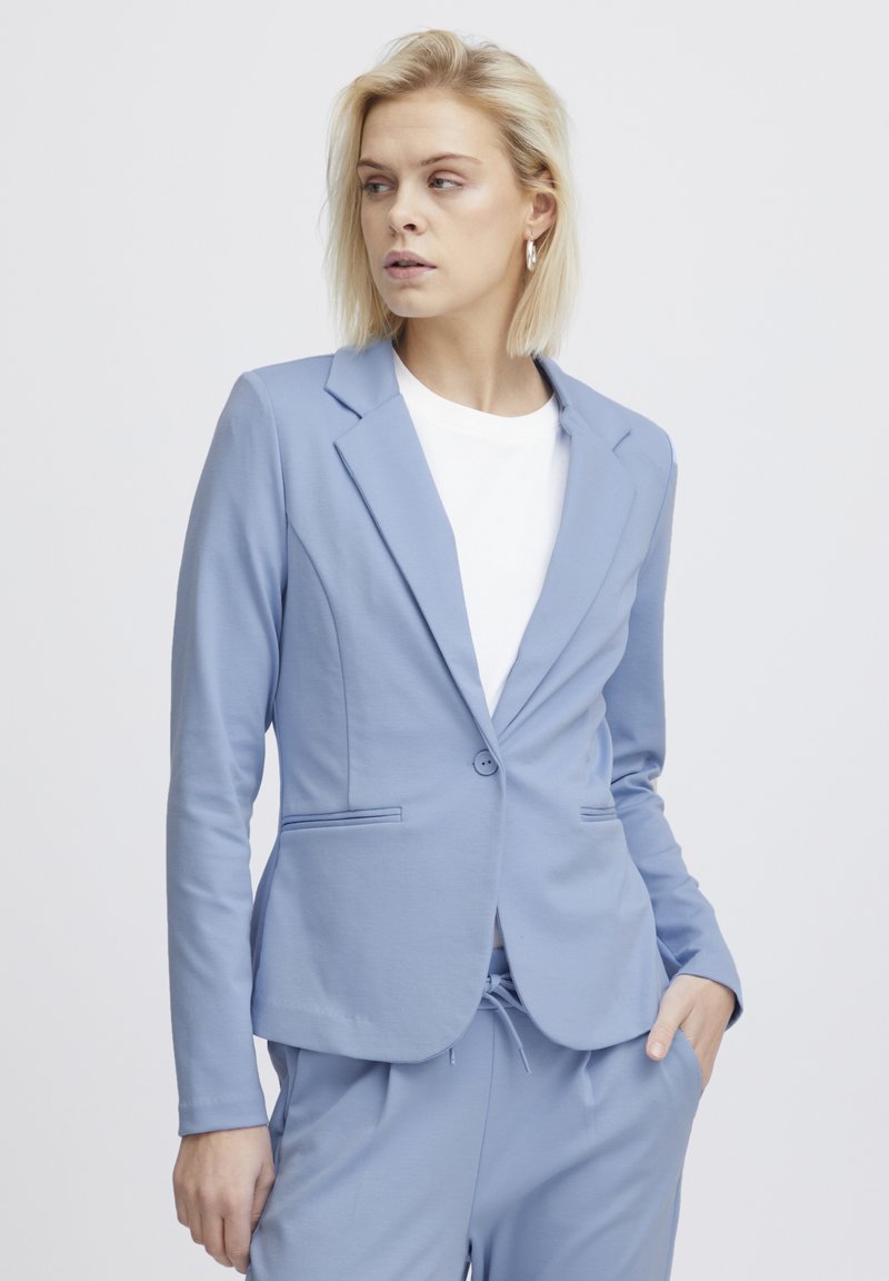 Lichtblauwe blazer van zacht fabric, met een enkele knoopsluiting, twee voorzakken en een getailleerde pasvorm, gecombineerd met een witte top.