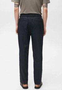 Diepblauwe op maat gemaakte broek met een elastische tailleband, zijzakken en een gladde, lichte stof; gedragen met een grijs shirt en sandalen.