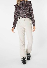 Mustapusero pitkillä hihoilla, kukkakuviolla ja röyhelöillä olkapäillä, yhdistettynä vaaleanbeigeihin housuihin, joissa on vetoketjulliset taskut ja tummat hiihtokengät.