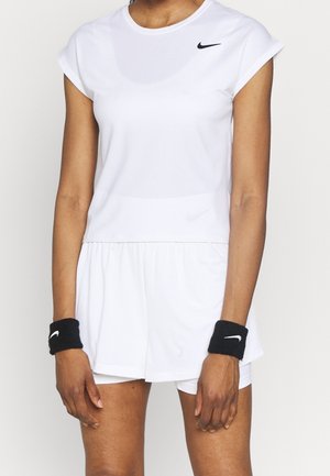 Femme portant un haut de sport blanc à manches courtes, une jupe plissée blanche, et des bracelets noirs avec des logos Nike blancs, debout devant un fond uni.