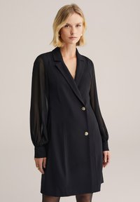 Rochie neagră cu un design ajustat, mâneci lungi din material transparent și accente cu nasturi aurii. Material neted cu o siluetă structurată.