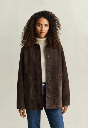 Frau mit lockigem Haar, die eine dunkelbraune, geknöpfte Wildlederjacke, einen weißen Rollkragenpullover und blaue Jeans trägt, steht vor einem schlichten beigen Hintergrund.