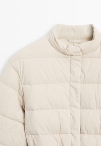 Giubbotto imbottito beige trapuntato con colletto rialzato e chiusura frontale a bottoni a pressione, mostrato dalla spalla al petto su sfondo bianco.