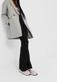 Grauer Wollmantel mit zwei vorderen Knöpfen, schwarzen, ausgestellten Hosen und helllila-grauen Sneakers mit schwarzen Sohlen und Schnürdetails.