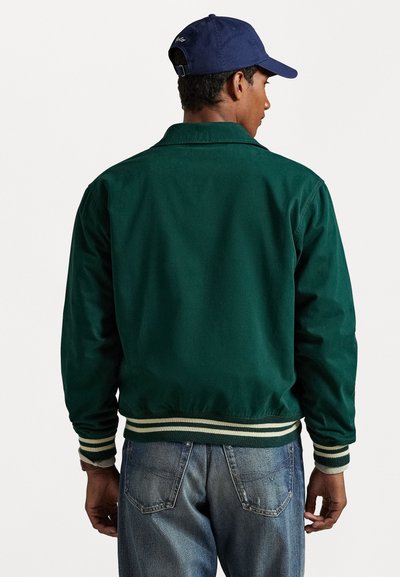 Veste bombardier verte avec un ourlet et des poignets côtelés rayés, dotée d'un col et d'un empiècement au dos, associée à un jean en denim bleu.