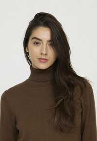 Pull en col roulé marron en tissu côtelé, avec des manches longues et une coupe ajustée, porté avec des cheveux lâchés et peu d'accessoires.