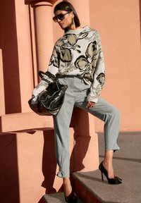 Vrouw in een oversized bloementrui, grijze op maat gemaakte broek, zwarte hoge hakken, grote zonnebril, en met een zwarte handtas bij een perzikkleurige muur.