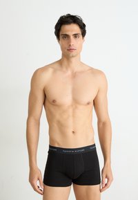 Fekete pamut boxer alsónadrág piros és fehér logós pánttal. A modellen atletikus testalkat van, sima fehér háttér előtt áll.