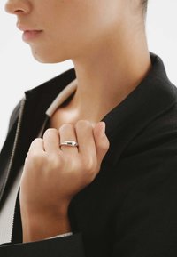 Anillo de plata con un diseño suave y redondeado, llevado en una mano que sostiene una chaqueta de tela negra; el fondo es de un color claro y neutro.