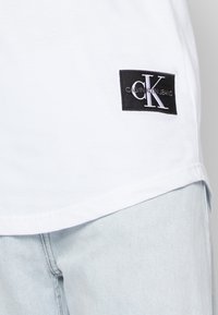 Biała bawełniana koszulka z czarną prostokątną naszywką z napisem "CALVIN KLEIN JEANS" oraz stylizowanym logo "CK". Gładka tekstura, luźny krój.