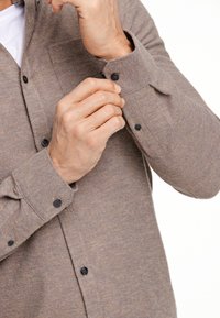 Camicia button-up marrone e texturizzata con bottoni neri. Primo piano di mani che regolano il polsino. Motivo sottile, maniche lunghe e taschino sul petto.