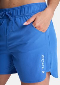 Μπλε αθλητικό σορτς με ελαστική μέση, πλαϊνή τσέπη και λευκό λογότυπο. Το ύφασμα φαίνεται λείο με ελαφρά γυαλάδα.