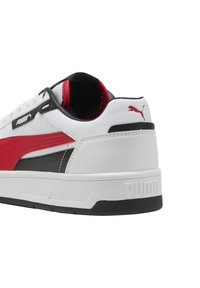 Sneaker bianco con accenti rossi e neri, texture in pelle, colletto imbottito e suola in gomma. Presenta un marchio Puma ben in evidenza sul tallone.