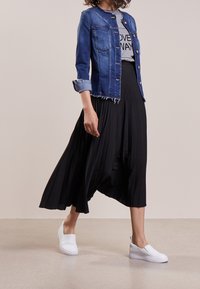 Jeansjacka med slitna kanter, grå grafisk t-shirt, flytande svart plisserad kjol och vita slip-on skor. Strukturerade material, avslappnad design.