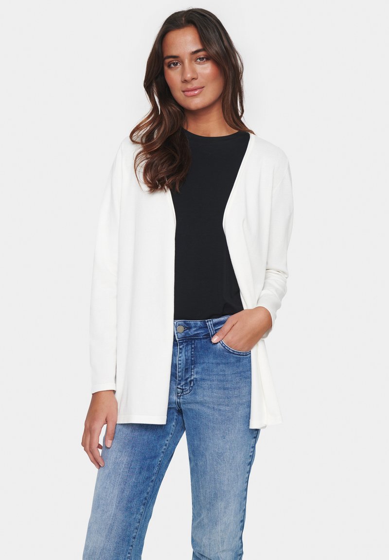 Cardigan bianco con apertura frontale, maniche lunghe e texture morbida, indossato sopra un top nero e jeans blu.