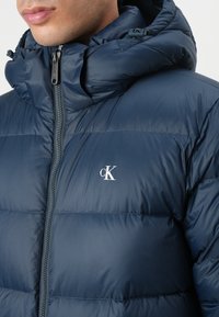 Marineblå puffjakke med høy krage, glidelås og et brodert hvitt logo på brystet. Har en strukturert, glanset overflate.