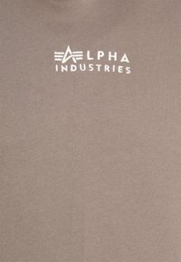 Béžové bavlnené tričko s bielym logom "ALPHA INDUSTRIES" v jednoduchom písmovom variante, s hladkou, jemnou textúrou a vo voľnom strihu.