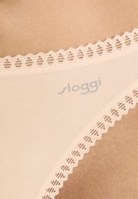 Beige Sloggi slip met een gladde textuur, kantafwerking met diamantvormige uitsparingen en een subtiele merklogo in lichtblauw.