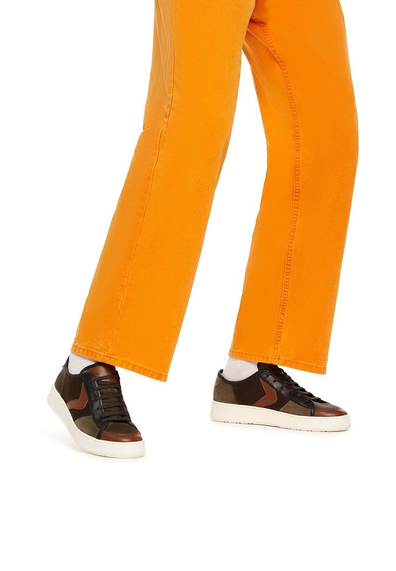 Jambes portant un pantalon orange vif, des chaussettes blanches et des baskets en cuir marron et noir avec semelles blanches sur fond blanc.