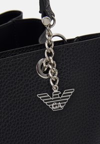 Czarna torebka skórzana o fakturze z metalowym łańcuszkiem w kolorze srebrnym oraz metalowym charmsie w kształcie orła z inicjałami "GA" przymocowanym w pobliżu uchwytu.