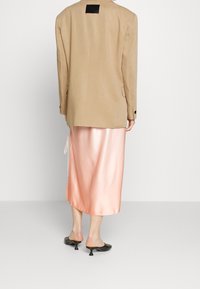 Blazer beige con una etiqueta negra cuadrada en la parte trasera, combinado con una falda de satén rosa clara, que presenta un detalle de lazo en la cintura. Mules negros de tacón.