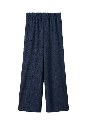 Mladá žena s dlhými vlasmi nosí tmavomodrý bezramienkový nariasený top, zodpovedajúce široké nohavice a čierne žabky na bielom pozadí.