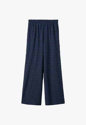 Jeune femme aux longs cheveux porte un top smocké bustier bleu marine, un pantalon large assorti et des tongs noires sur fond blanc.