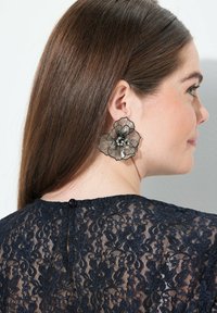 Orecchini neri a fiori con un design testurizzato, caratterizzati da linee delicate e piccoli accenti simili a perle, indossati con un top in pizzo.