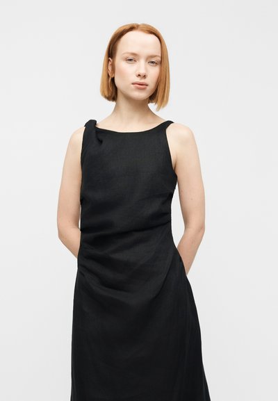 Μαύρο λινό φόρεμα με στρογγυλή λαιμόκοψη και λεπτομέρεια κόμπου στο μπροστινό μέρος. Το ύφασμα έχει ελαφριά υφή και πέφτει απαλά πάνω στη σιλουέτα.