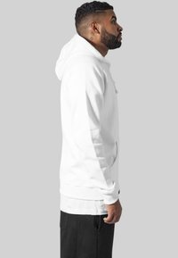 Witte hoodie met een kangoeroezak, verstelbare trekkoordkap, ribgebreide boorden en zoom; gecombineerd met zwarte broek, gezien vanaf de zijkant.